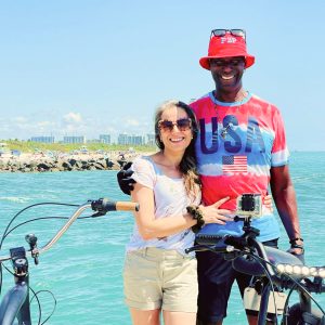 A man an a woman in front on a Bay. Man's arm is around the woman, and woman is holding the man's waist. Both are smiling. Bicycles are visible in the foregrand